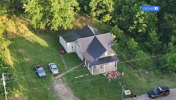 An aerial view of a residential property featuring a single-story house with a gray roof and a driveway lined by a few parked ve
