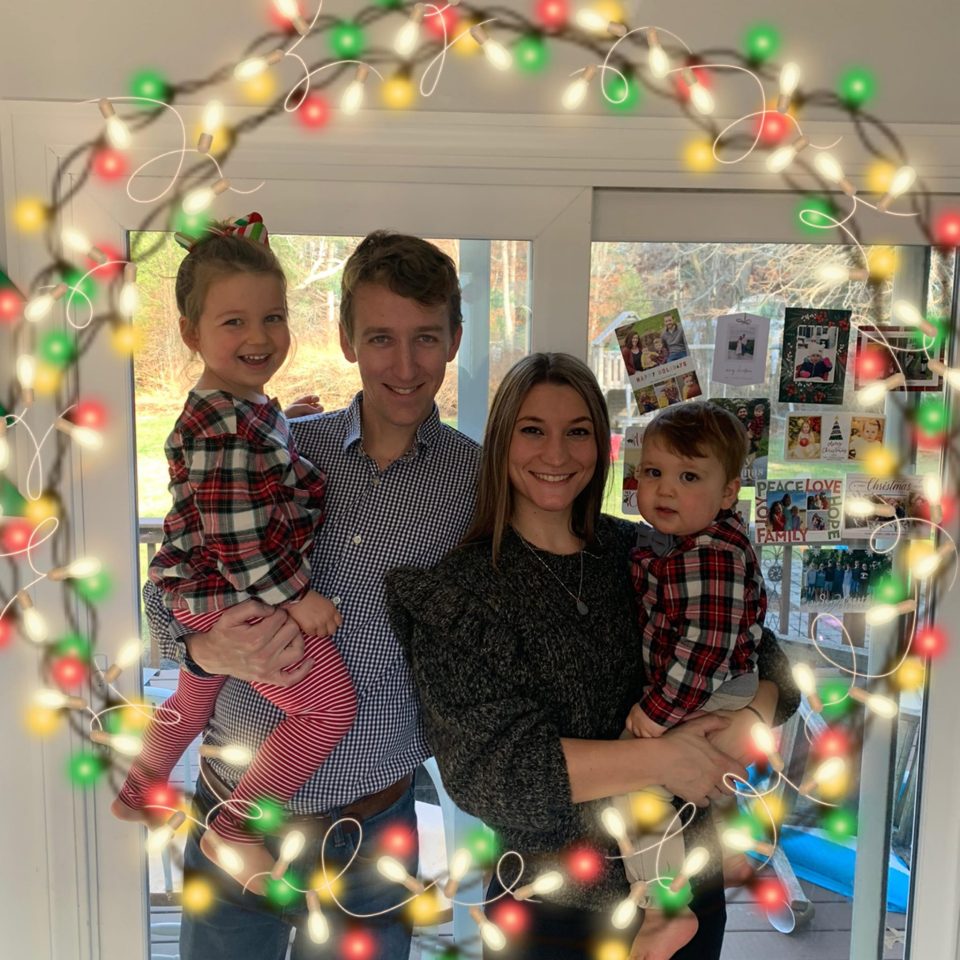 A family of four, including two young children, stands framed by Christmas lights.