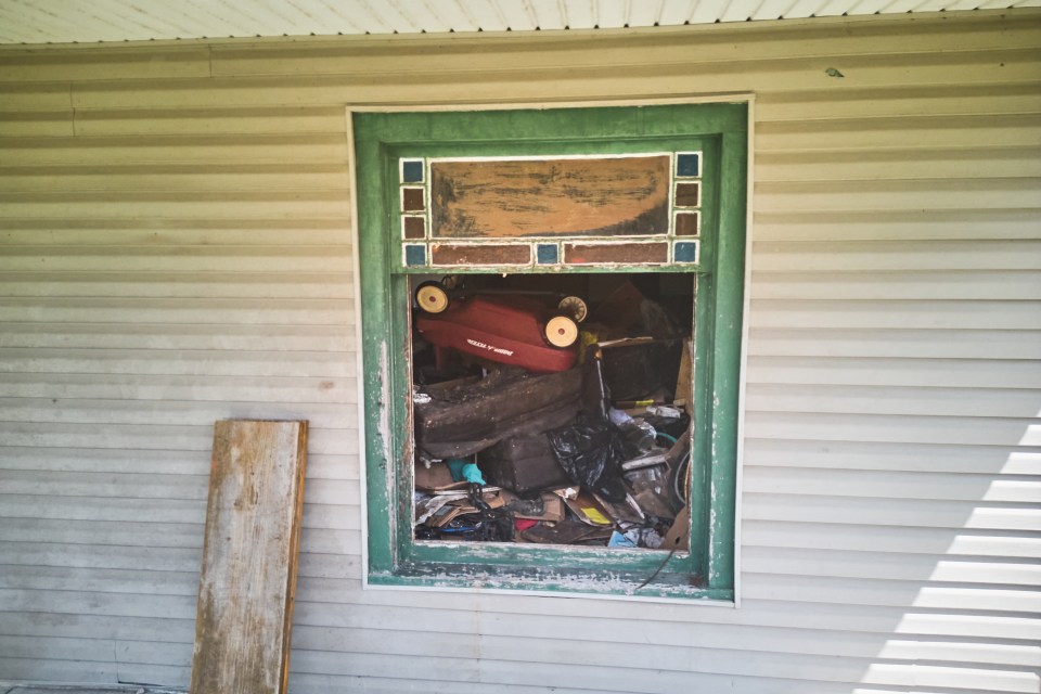 Debris, including a red wagon and black trash bags, fills the window frame of a home with white siding.