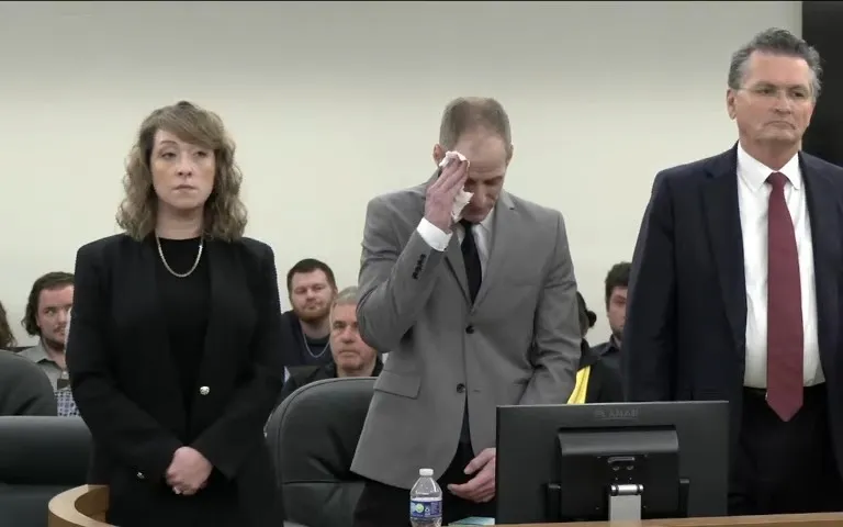 Three people stand in a courtroom, with the center figure wiping a tear.