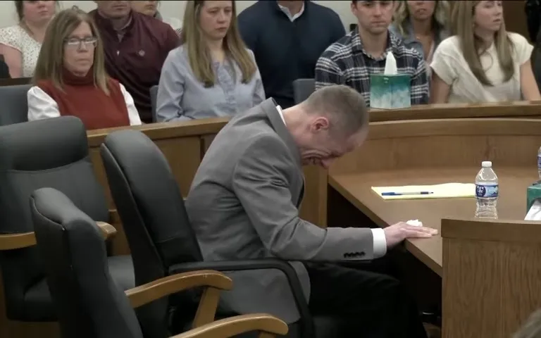 A man in a gray suit sits in a courtroom, leaning forward and reacting with emotion, while others watch from the gallery behind him.