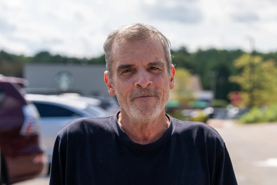 Eric McDonald, a man with graying hair and beard, wearing a dark blue t-shirt, looks directly at the viewer outdoors.