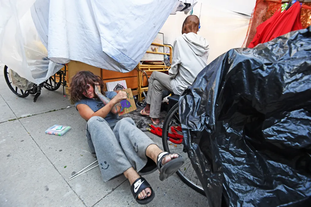 Barbara Andre sits on the ground with a canvas and art supplies next to an encampment of white tarps and a person in a white hoodie sitting on a chair.
