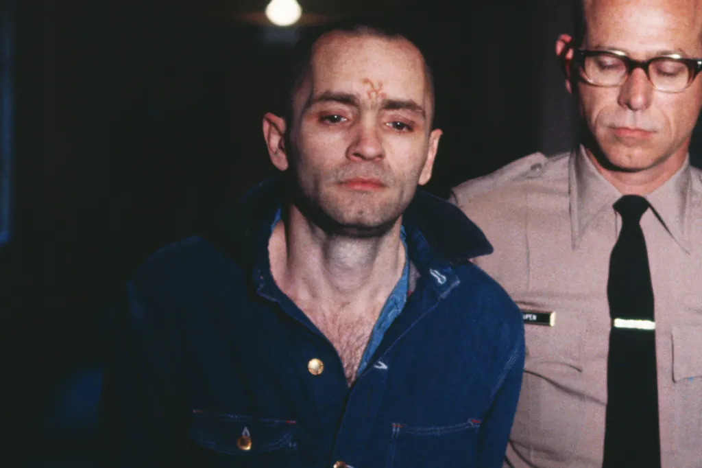A policeman escorting Charles Manson with an 'X' carved into his forehead.