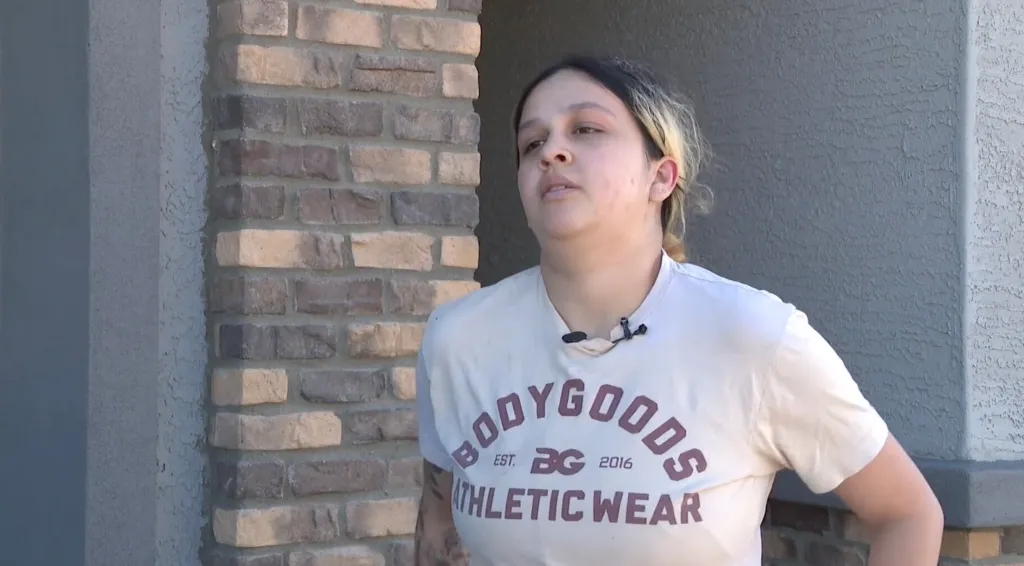 Bella Torres stands outdoors near a brick wall, wearing a light-colored t-shirt with a microphone clipped to the collar.