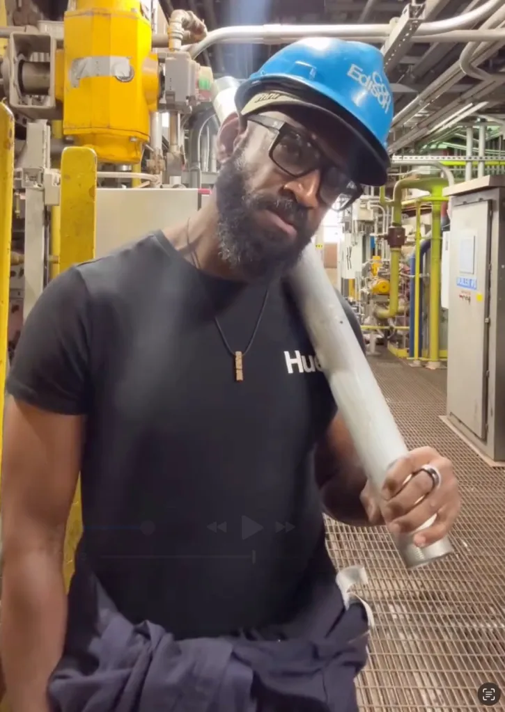 Koran Irvine wearing a hard hat and glasses while holding a pipe in an industrial setting.