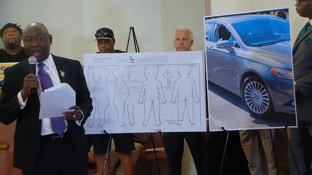 A speaker holds a microphone and documents in front of two display boards, one showing anatomical diagrams and the other a car with a shattered window.