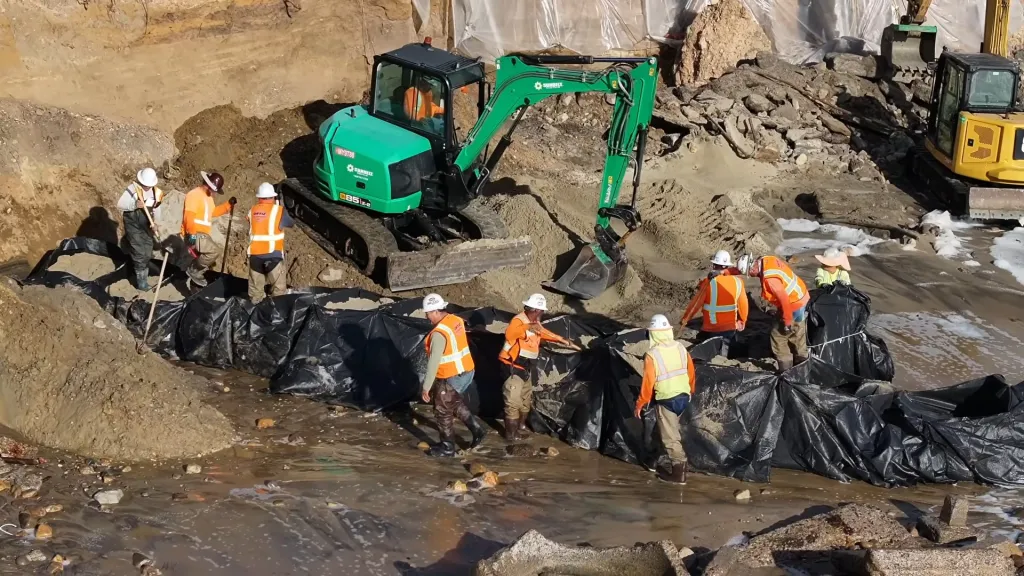 Construction workers in hard hats and safety vests fill large black bags with sand next to heavy machinery on a beach.