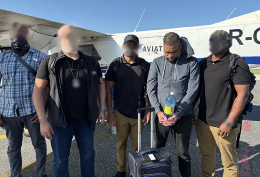 Kanand Ojha, flanked by ICE officers, stands in front of a plane with a suitcase and a water bottle.