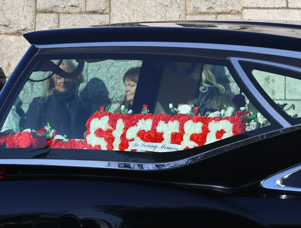Mourners seated inside a hearse beside a floral tribute that reads "SISTER".