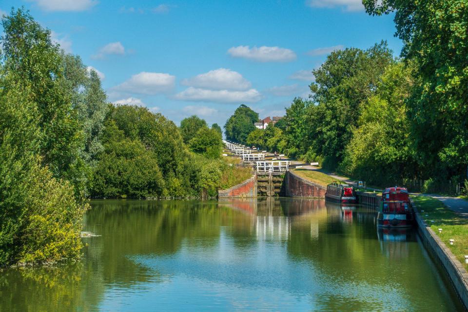 A view of the Caen Hill Lock Flight on the Kennet and Avon Canal, showing a series of canal locks rising up a lush green hill under a blue sky.