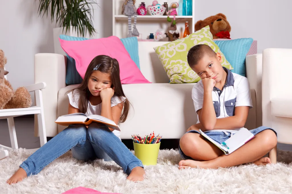 Two bored children sitting on the floor in front of a sofa, holding books and resting their chins on their hands.