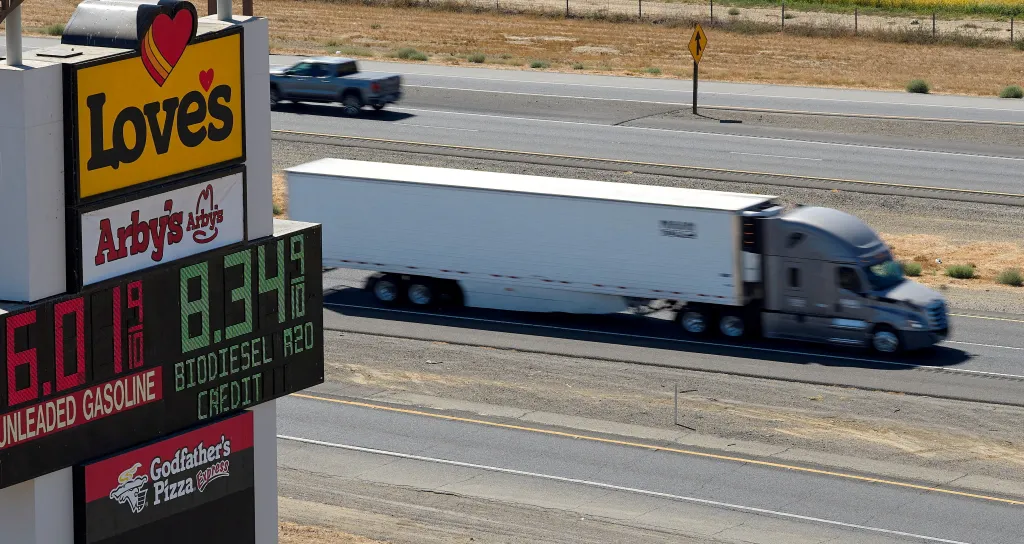 A semi-truck drives past a highway sign displaying fuel prices.