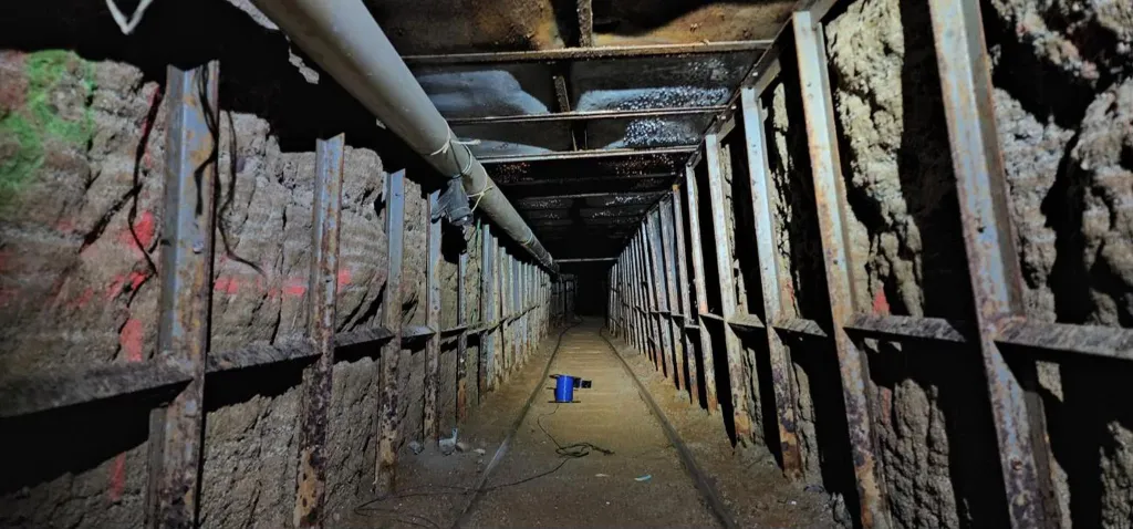 The interior of a cross-border smuggling tunnel, featuring reinforced walls, a ventilation pipe, and a rail system stretching into the distance.