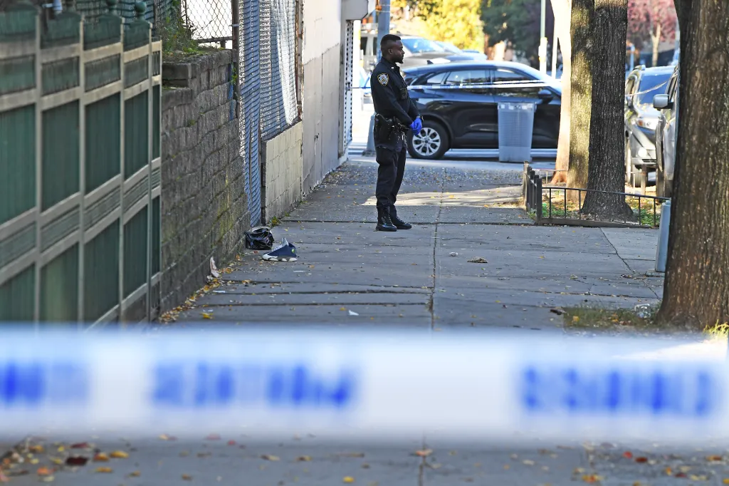 An NYPD officer stands guard at a Bronx shooting scene near Crotona Park, marked by police tape across the foreground and background.