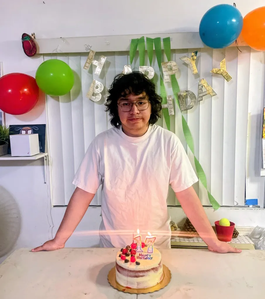 A young man in glasses and a white t-shirt stands behind a 17th birthday cake with lit candles, surrounded by colorful balloons and birthday decorations.