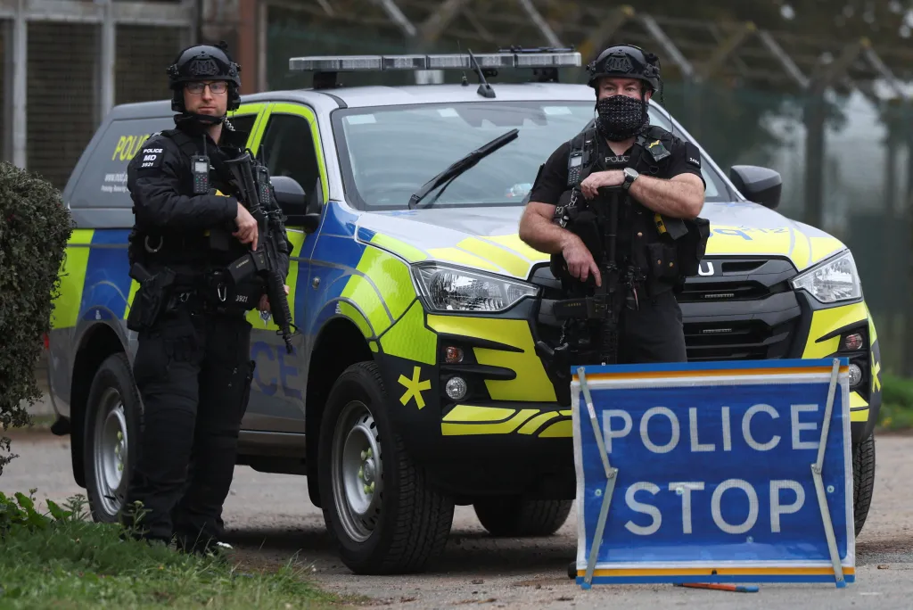 Two armed police officers stand in front of a police vehicle and a "Police Stop" sign near the RAF Fairford airbase.