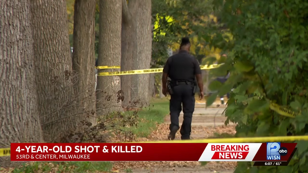 A police officer walks along a sidewalk lined with trees, surrounded by yellow police tape, with a news graphic at the bottom reading "4-Year-Old Shot & Killed."
