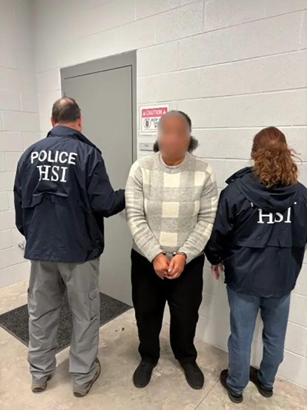 Marva Cook flanked by two HSI agents in handcuffs.
