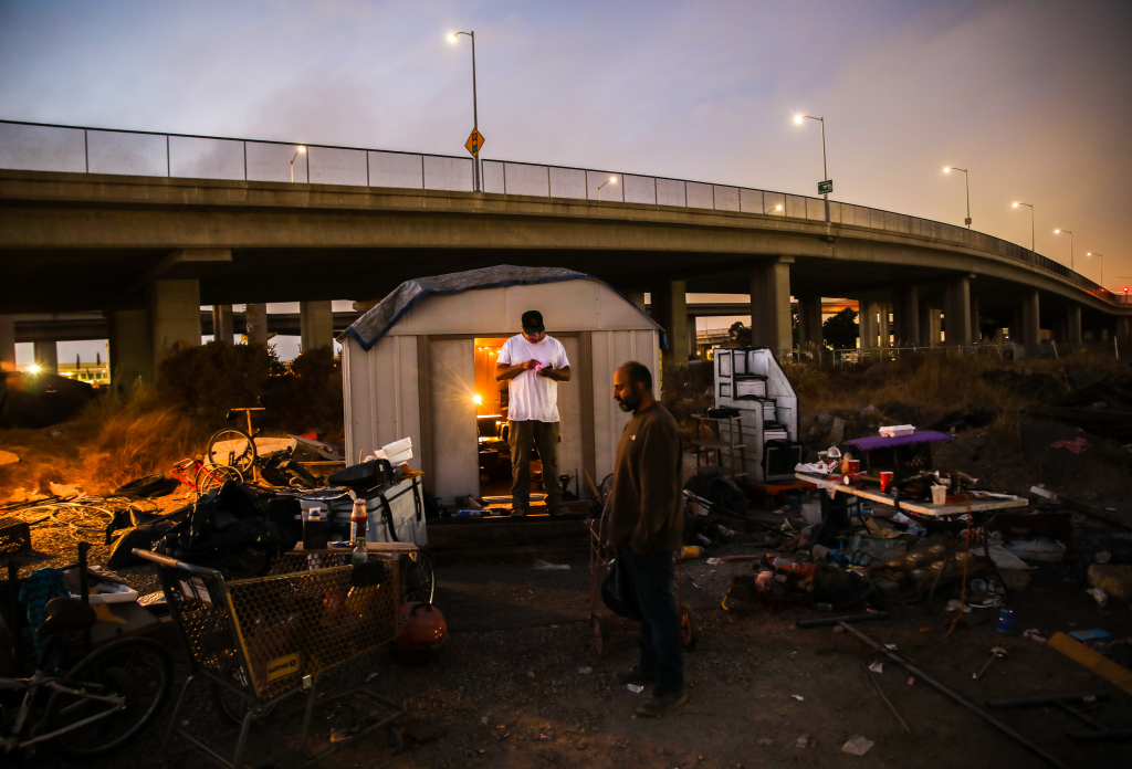 Gabriel Cano and Moose Saberi relax outside Saberi's shed at an encampment under a highway overpass in Oakland.