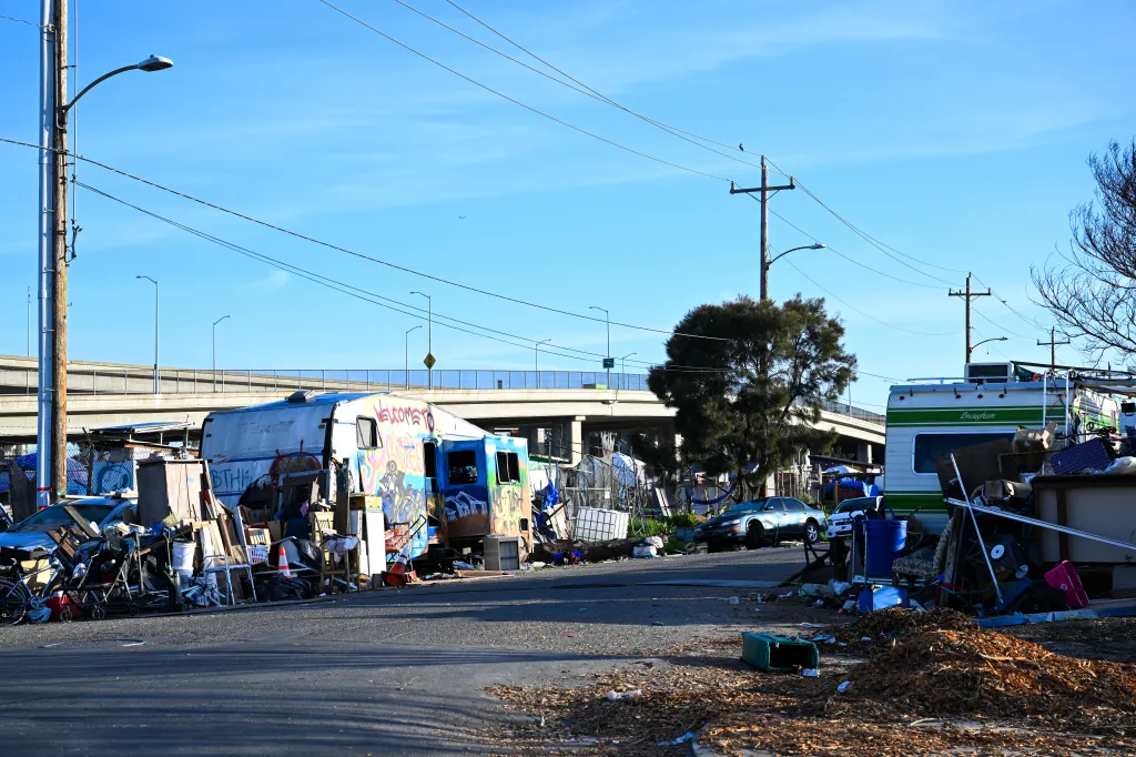 A homeless encampment lines a street in West Oakland, California, with RVs, makeshift shelters, and debris scattered along the roadside.