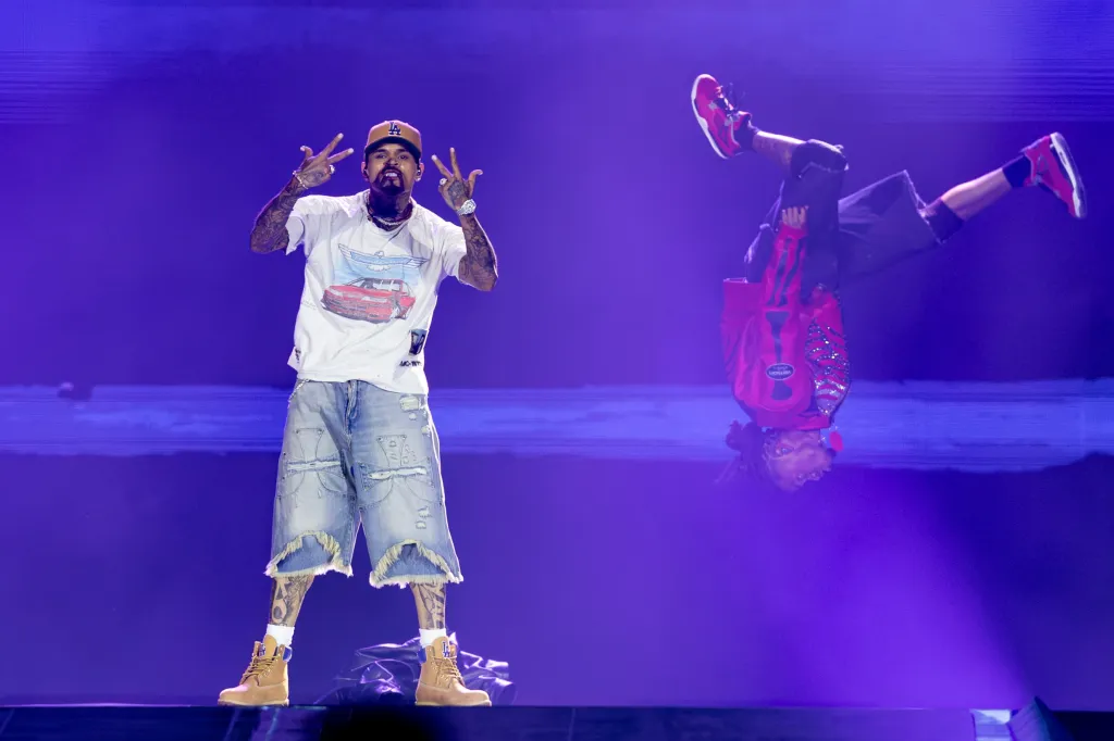 Chris Brown performs on stage, flashing hand signs while another performer does a backflip mid-air.