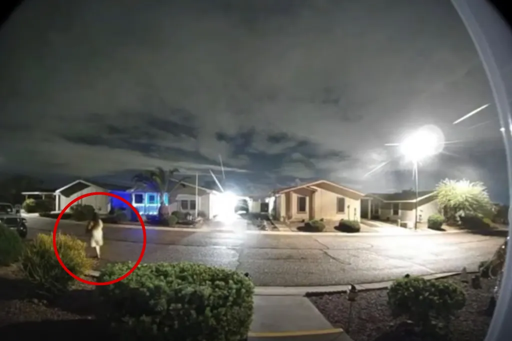 A neighborhood street at night captured on a security camera, featuring a figure circled in red walking down the sidewalk.