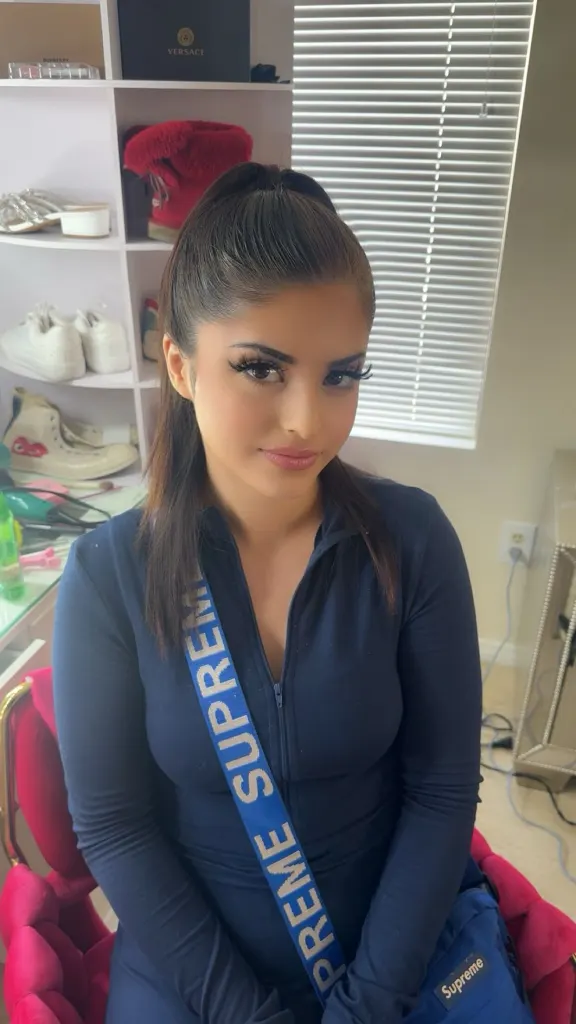 Mina Kurdian indoors, wearing a dark blue zip-up top with a blue "Supreme" strap across her chest, with shelves containing shoes and boxes in the background.