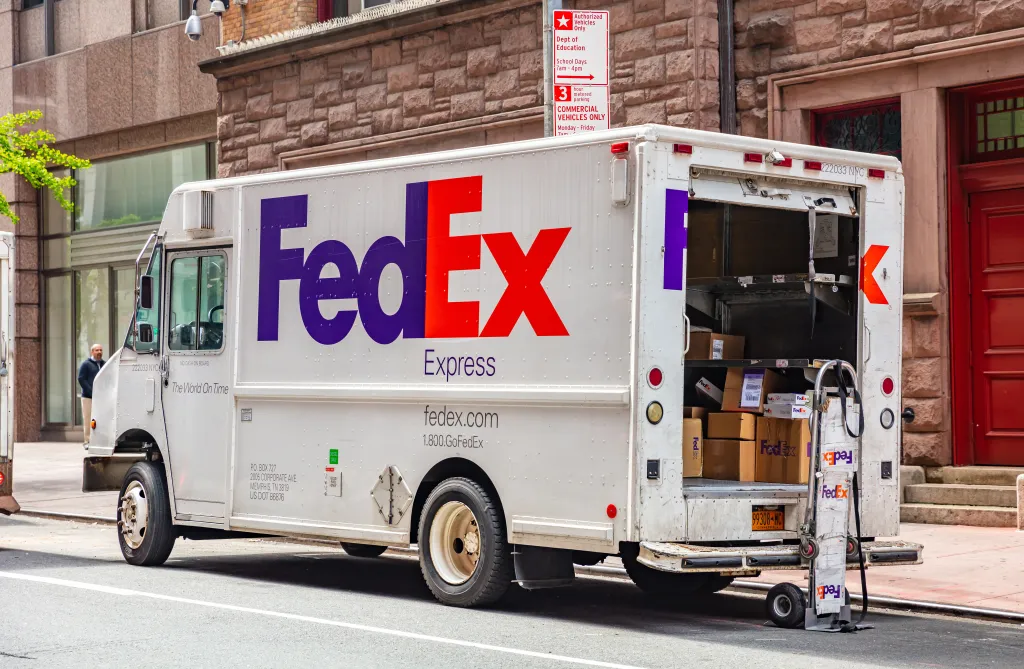 A FedEx delivery truck is parked on a city street with its rear door open, revealing boxes and a hand truck inside.