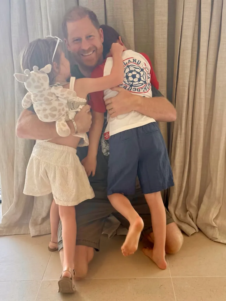 Prince Harry kneels and smiles while embracing his young children, Prince Archie and Princess Lilibet.