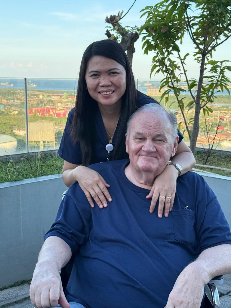 Thomas Markle in a blue shirt sitting in a wheelchair with a nurse standing behind him on a rooftop overlooking a city.