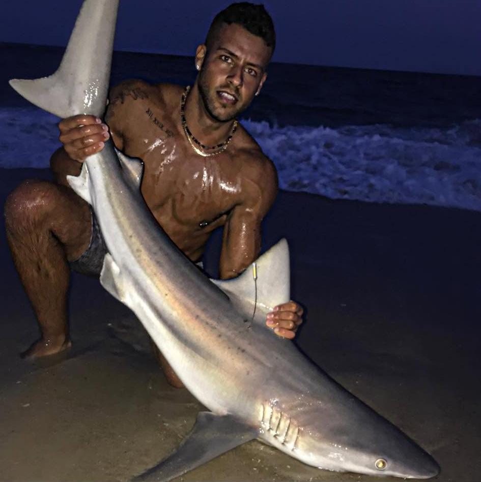 A shirtless man posing on a beach at night, holding a large shark.