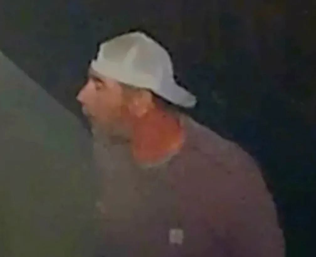 Surveillance footage of a man wearing a light-colored baseball cap looking through a window.