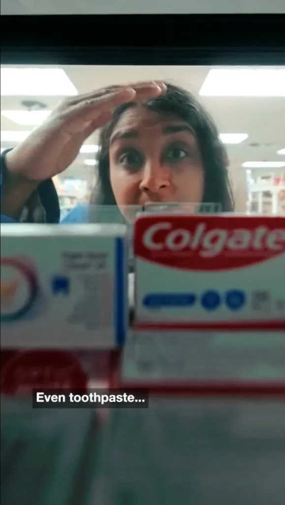 A person peers through the glass of a locked retail store shelf containing boxes of Colgate toothpaste, with the caption "Even toothpaste...".