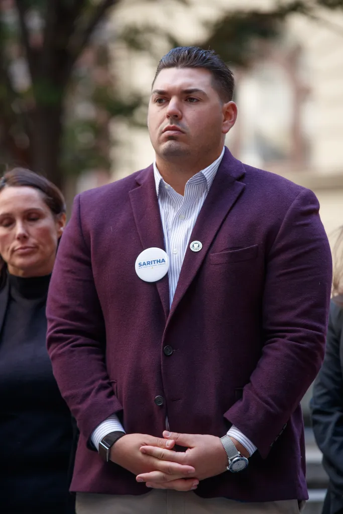 A man wearing a maroon blazer and a Saritha Komatireddy campaign button stands with his hands clasped at a press conference.