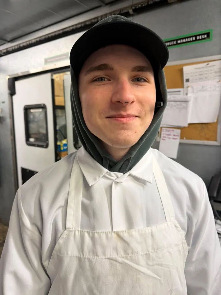 Portrait of Maclaine Schmidt wearing a white chef's coat, apron, cap, and a dark hoodie.