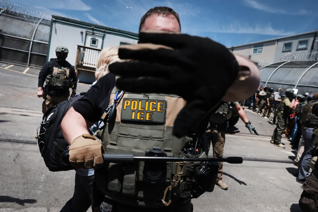 An ICE officer wearing tactical gear blocks the camera lens with a gloved hand outside a detention facility, with protesters visible in the background.
