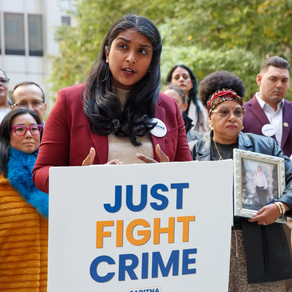 New York Attorney General candidate Saritha Komatireddy speaks at a press conference behind a podium that reads "JUST FIGHT CRIME," flanked by family members holding a framed photo of a loved one.