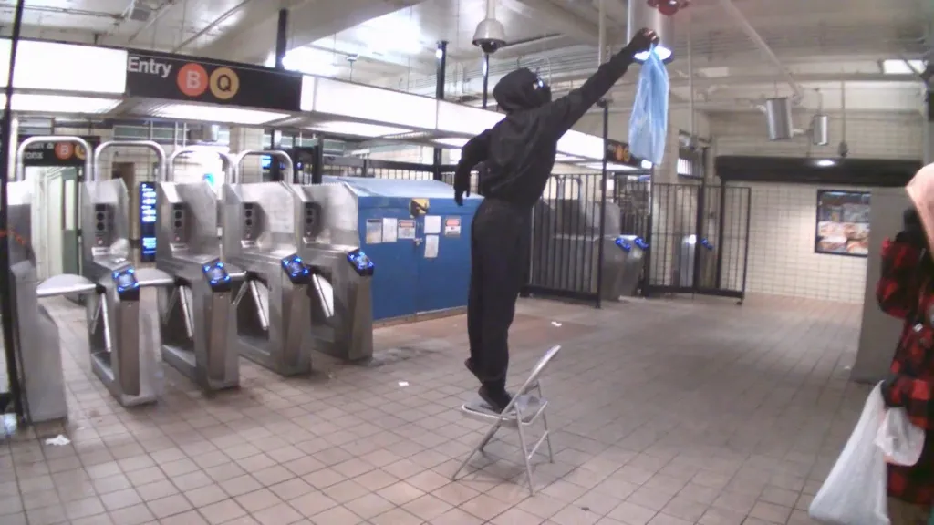 A masked suspect stands on a folding chair to cover a security camera in a subway station.