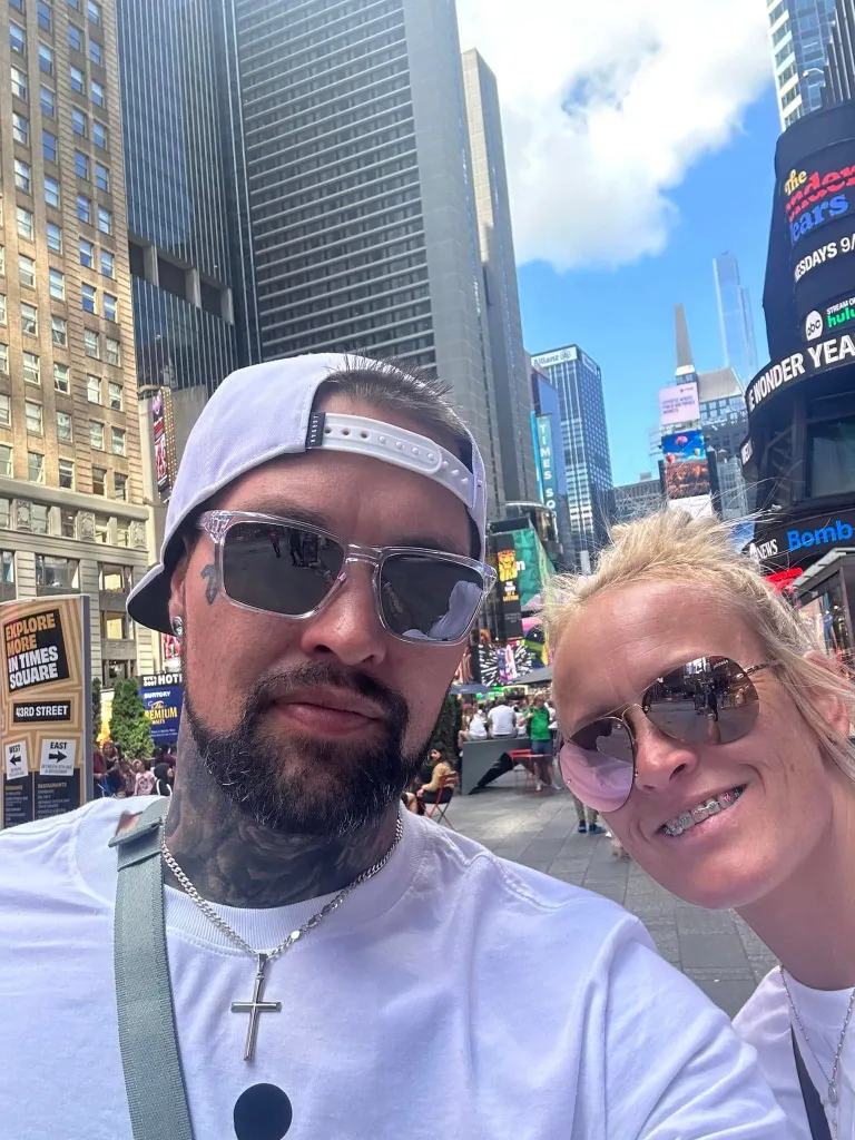 A man and woman taking a selfie in Times Square, New York City, surrounded by tall buildings and billboards.