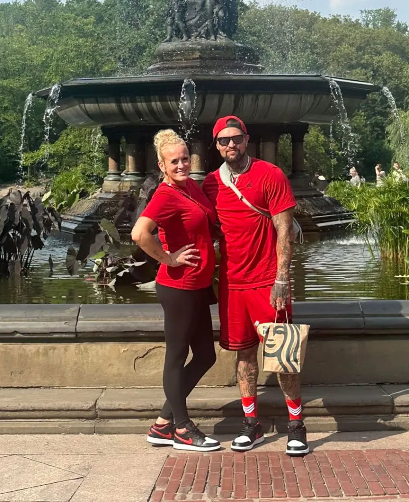 Cody and Honey Phipps pose in front of a fountain in a park, both wearing matching red tops and black-and-red sneakers.