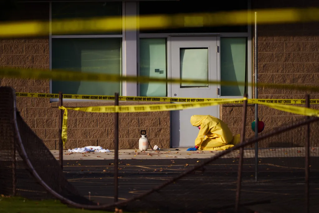 A person in a yellow hazmat suit cleans the crime scene outside an elementary school behind yellow police tape.