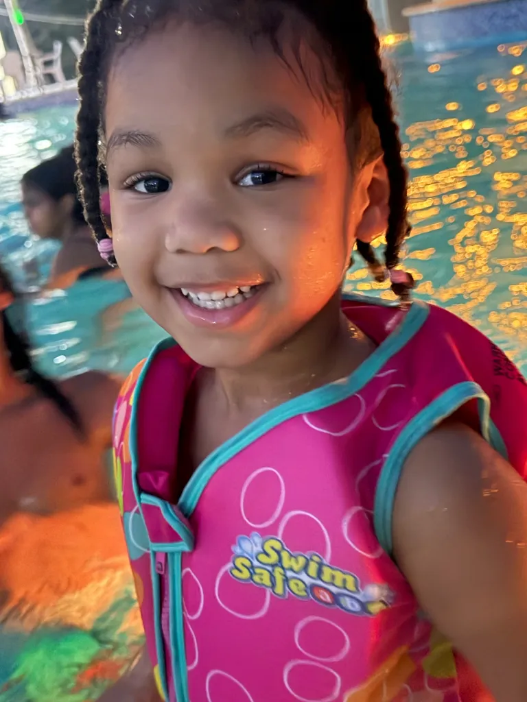 A young child wearing a pink and blue life vest smiles in a swimming pool.