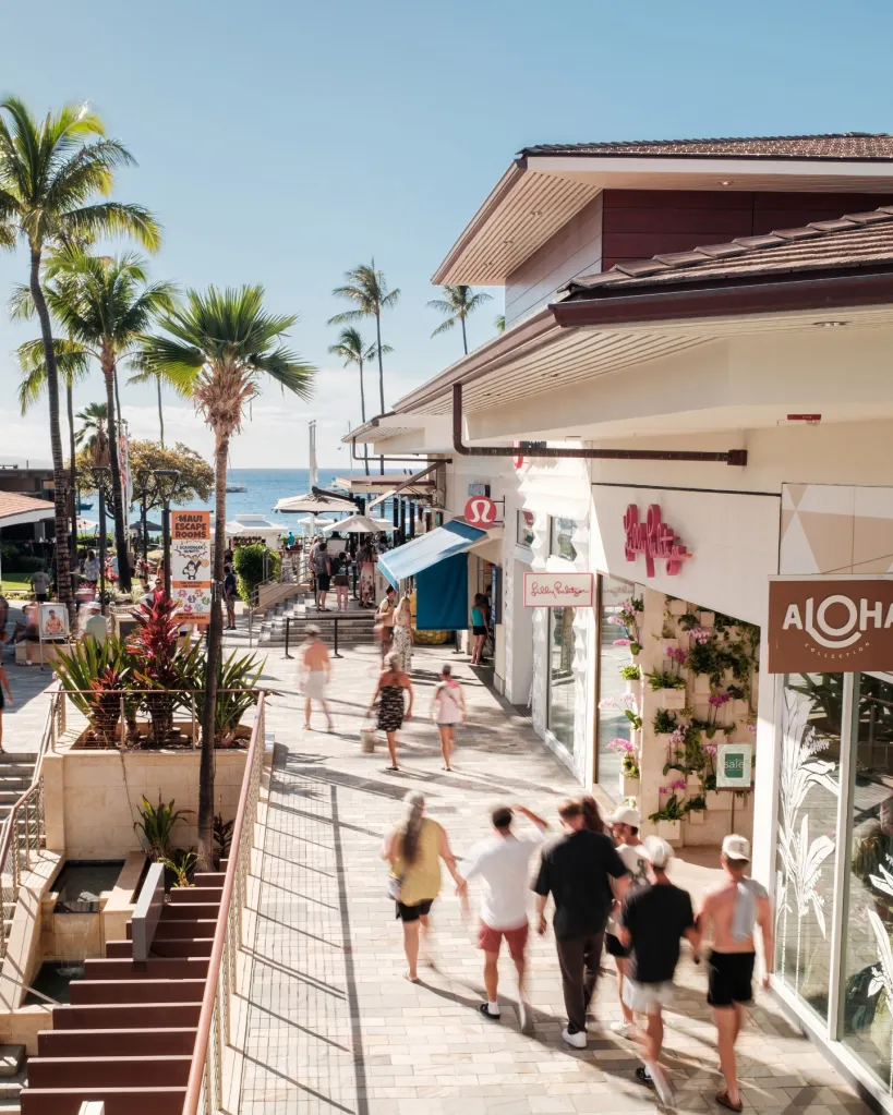 People walking along the sunny outdoor shopping corridor at Whalers Village in Kaanapali, flanked by palm trees and ocean views.