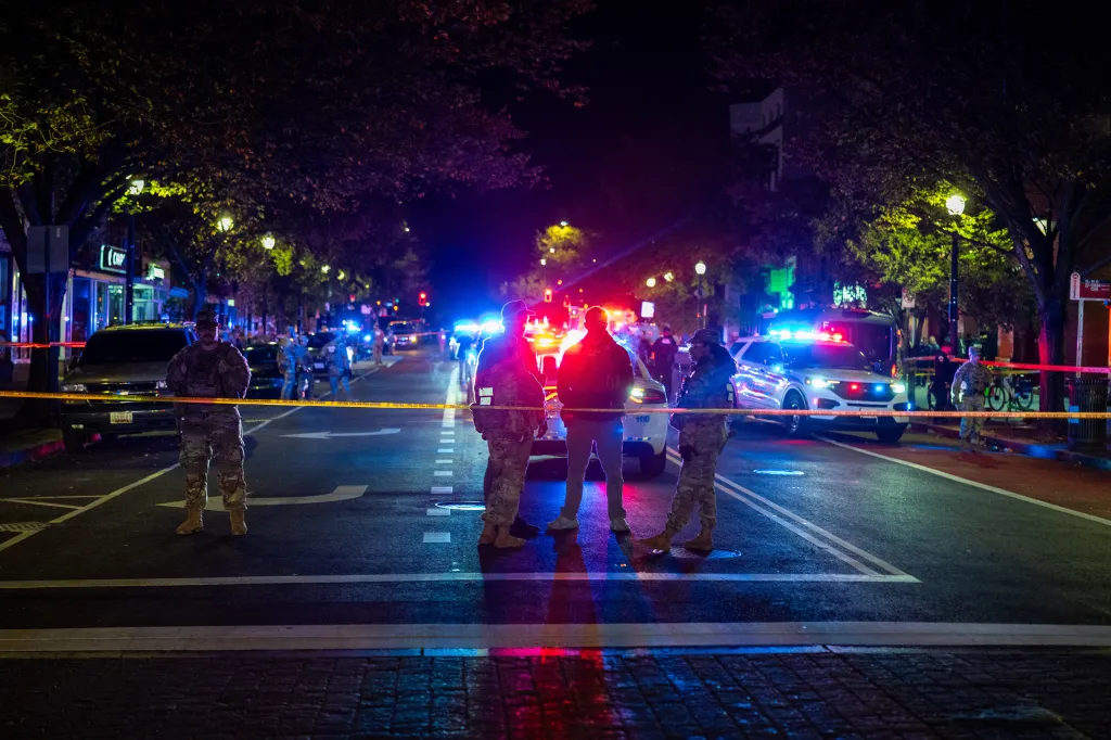 National Guard soldiers stand behind police tape at a cordoned-off street illuminated by emergency vehicle lights at night.
