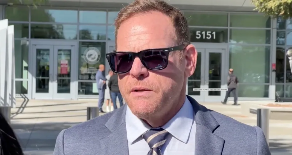 A man wearing a grey suit, light-blue shirt, striped tie, and sunglasses stands outside a building with the number 5151 on it.